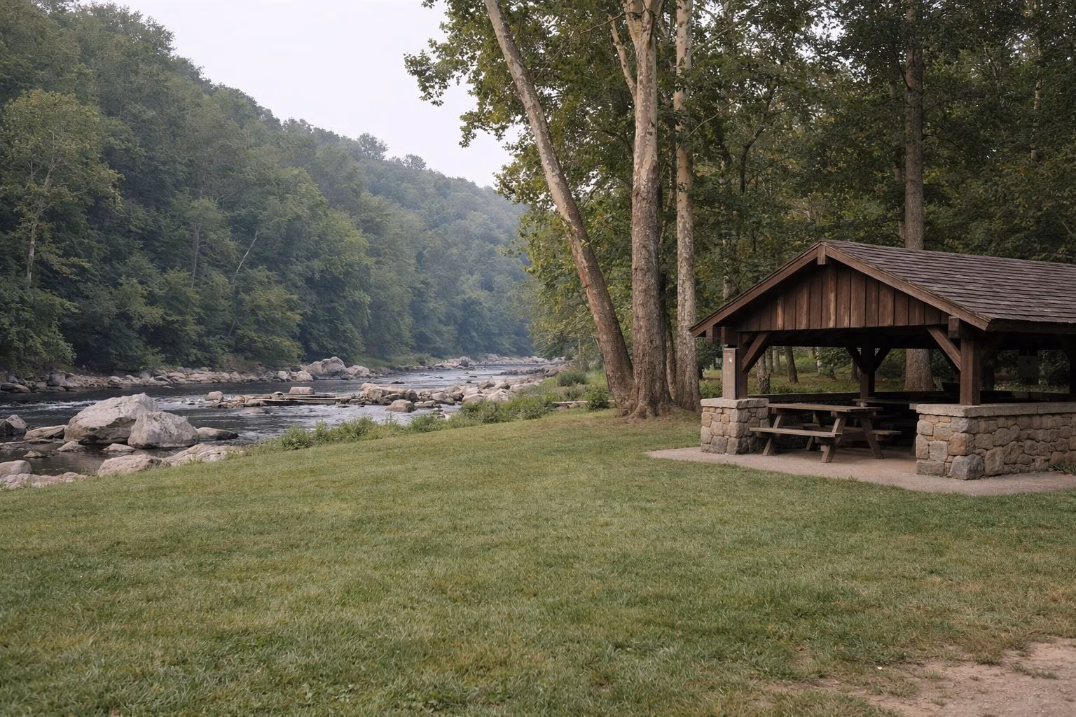 A mown picnic lawn under mature sycamores beside a wide shallow river, a stone-and-timber picnic shelter at the edge of the lawn