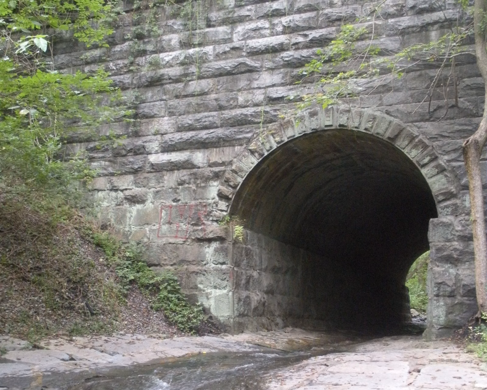 A stone-arched tunnel under the railroad along the Grist Mill Trail, a shallow stream running over flat rock in front of it