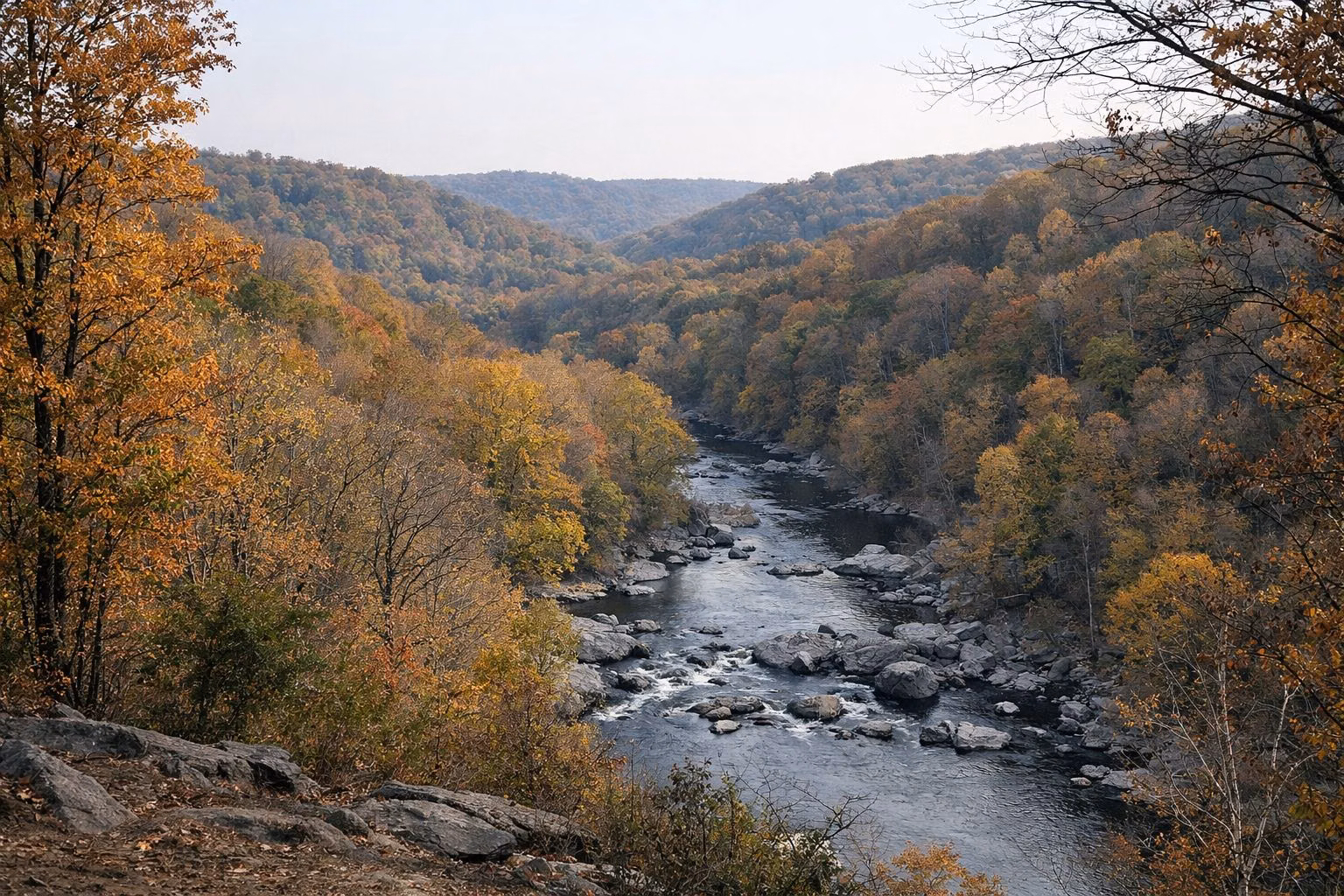 View from a hillside overlook across a forested river valley in peak fall color, layered wooded ridges, river glinting through the trees
