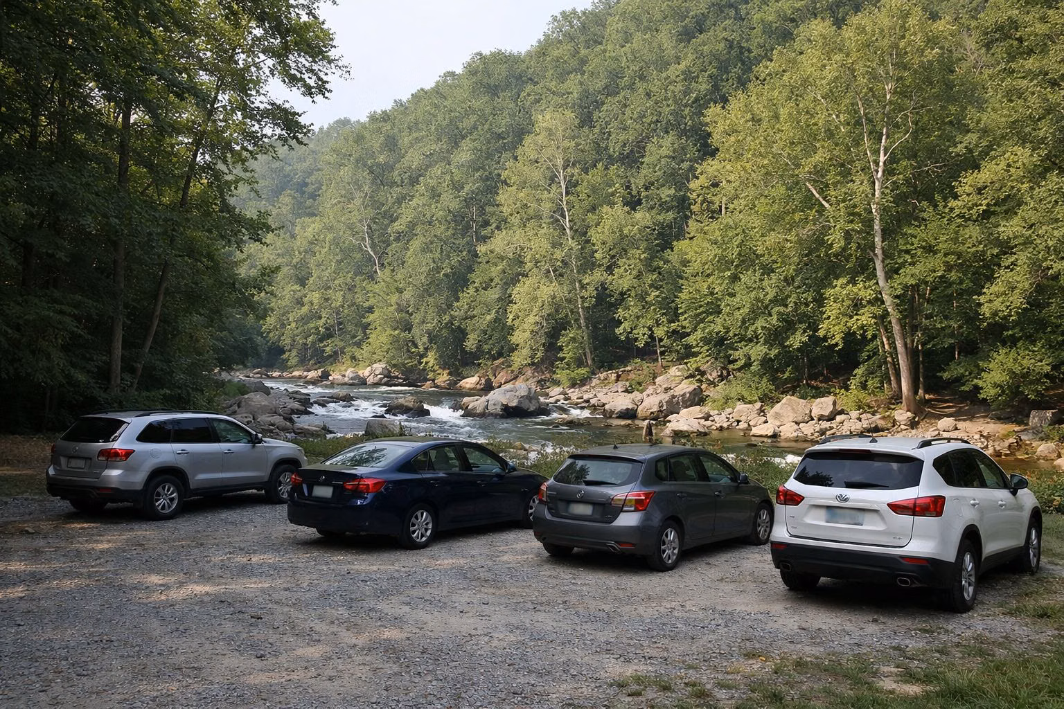 A small gravel parking lot at a wooded trailhead with a few cars parked, trees all around, no readable license plates