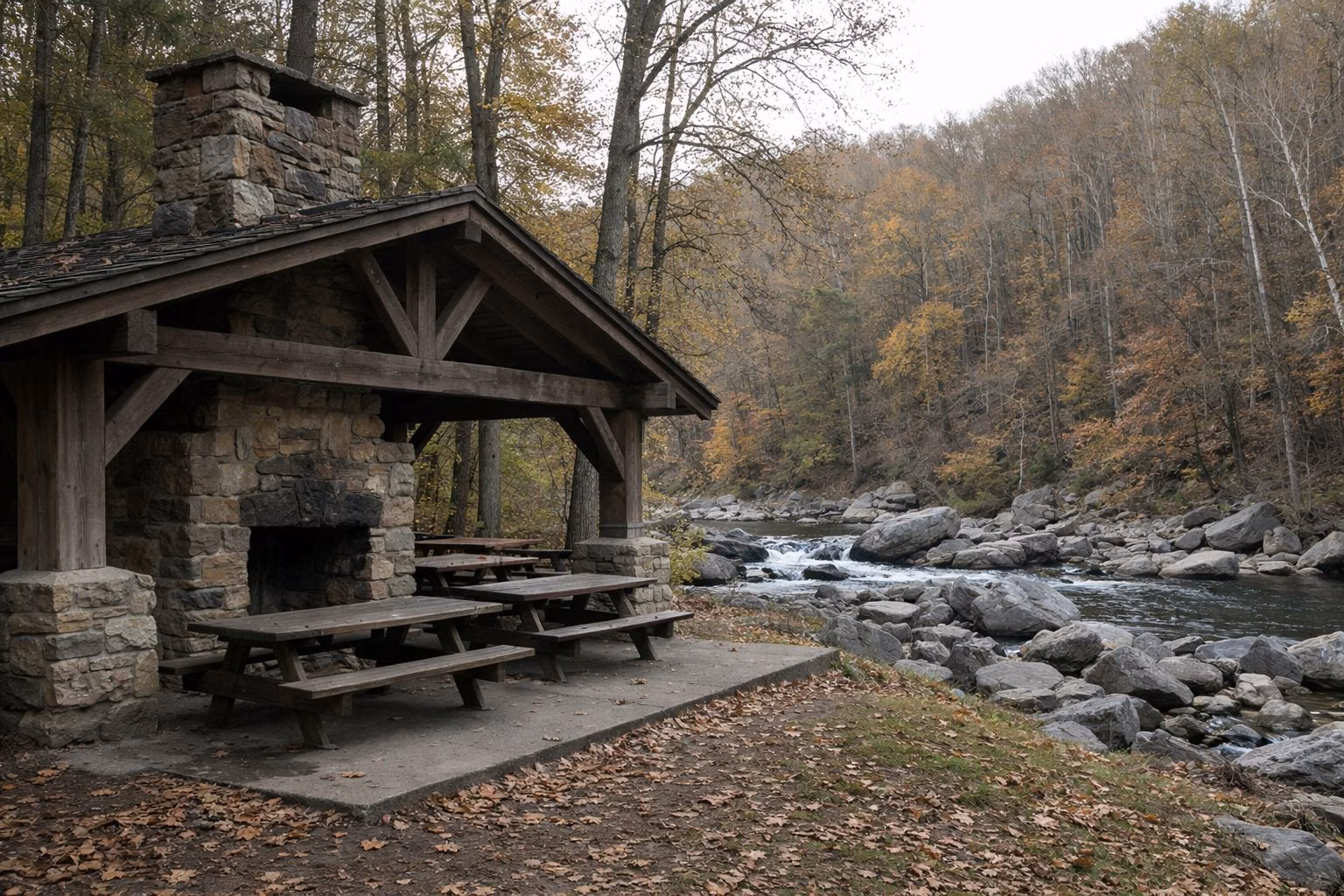 A rustic cut-stone and heavy-timber picnic shelter with a stone fireplace chimney, set in a hardwood forest, picnic tables inside
