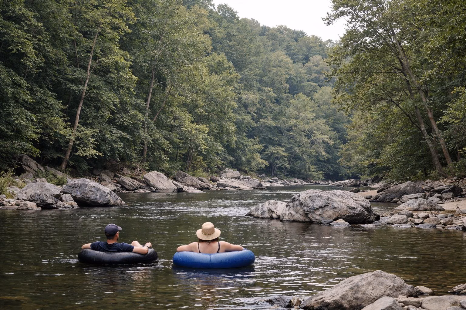 Two people floating on inner tubes down a calm stretch of a wooded river, small in the frame, seen from behind