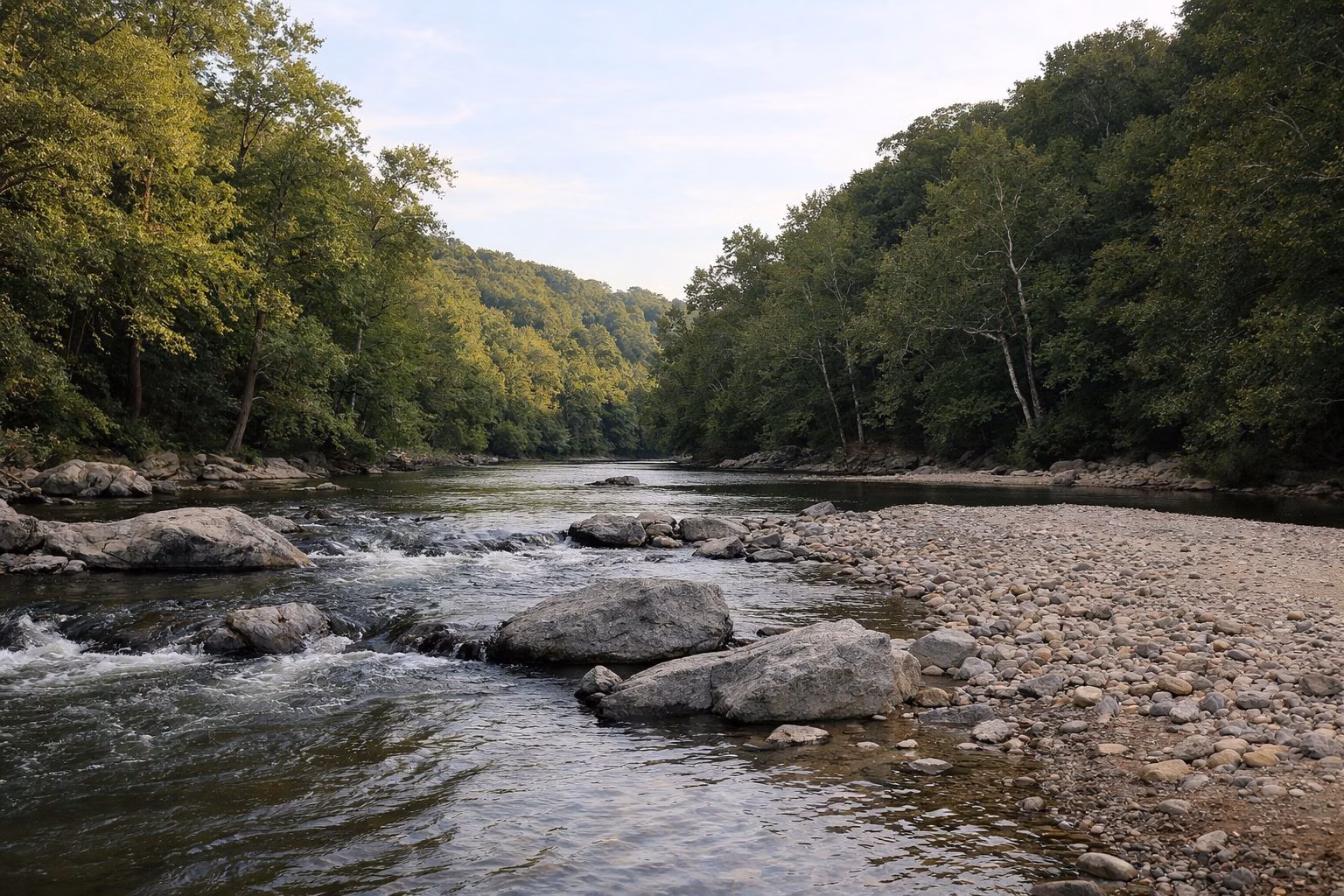 A shallow rocky river with a gravel bar and riffles, wooded hills rising on both banks, warm low sunlight