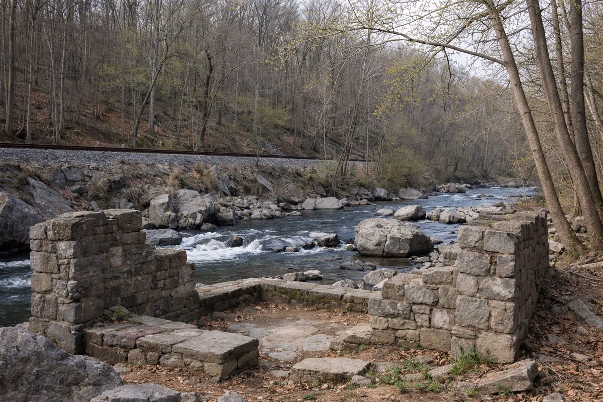 Old cut-stone foundation walls on a wooded riverbank below a railroad embankment, bare trees with first green buds